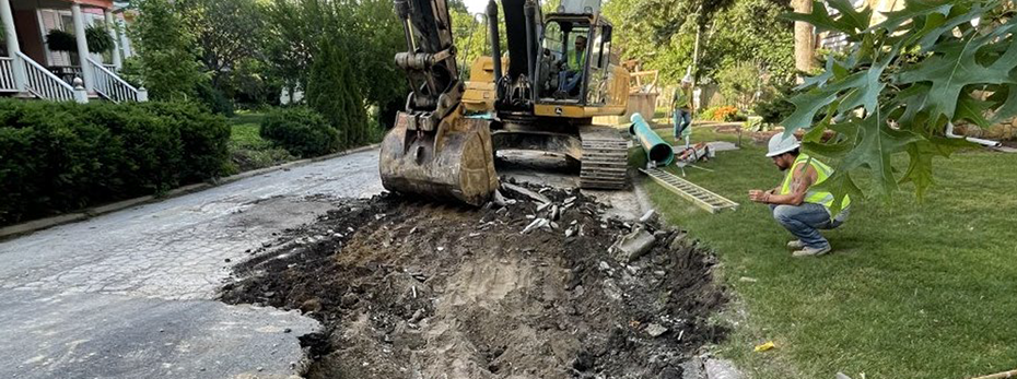 Worker in large backhoe removing asphalt from a road with workers and tools around it
