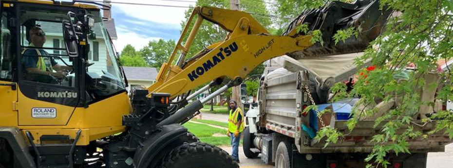 A person uses an end loader to load a mattress into a dump truck while another person observes