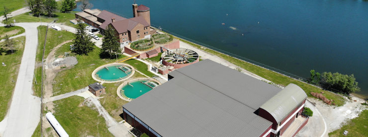 A drone photo of a water treatment plant with grass surrounding it and one of the shores of Lake Bloomington on the right side. It includes multiple buildings, round pools of water, and other pieces of equipment for treating water.
